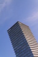A tall modern skyscraper with a distinctive tiered design, featuring glass and steel architecture, set against a clear blue sky. The building displays a prominent corporate logo at the top.
