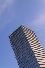 A tall modern skyscraper with a distinctive tiered design, featuring glass and steel architecture, set against a clear blue sky. The building displays a prominent corporate logo at the top.