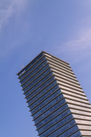 A tall modern skyscraper with a distinctive tiered design, featuring glass and steel architecture, set against a clear blue sky. The building displays a prominent corporate logo at the top.
