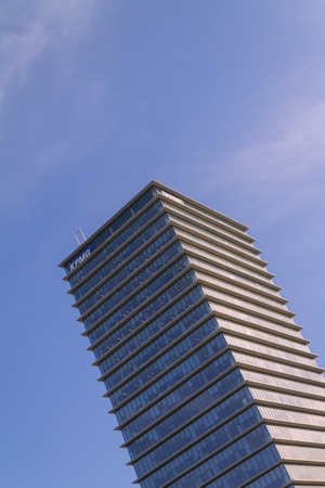 A tall modern skyscraper with a distinctive tiered design, featuring glass and steel architecture, set against a clear blue sky. The building displays a prominent corporate logo at the top.