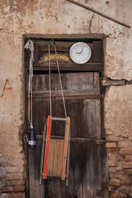 An aged, wooden door with a hanging wooden chair and a blue bottle tied with rope. Above the door, a sign with 'ALBERTAS ♥ RITA' is displayed alongside a round wall clock showing ten minutes past ten. The surrounding wall is weathered and textured with cracks.