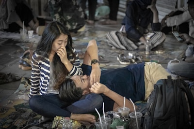 A young couple is sitting on a busy street floor. The woman, wearing a striped shirt, is sitting up while the man is lying with his head on her lap. There are food and drink containers around them, indicating casual dining or street food. Other people are visible in the background, some engaged with their phones.