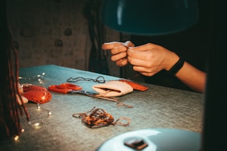 Hands engaging in a sewing or crafting activity on a table, with needles and thread visible. There are red scissors and small stitched objects, possibly made of felt. Soft lighting casts a warm, focused atmosphere.