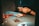 Hands engaging in a sewing or crafting activity on a table, with needles and thread visible. There are red scissors and small stitched objects, possibly made of felt. Soft lighting casts a warm, focused atmosphere.