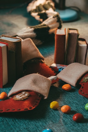 A collection of small, homemade felt pouches in earthy tones is scattered on a textured surface. They are accompanied by colorful candy-coated chocolates and partially opened candy wrappers. In the background, there is a cozy knit scarf and a blurred, illuminated lamp.