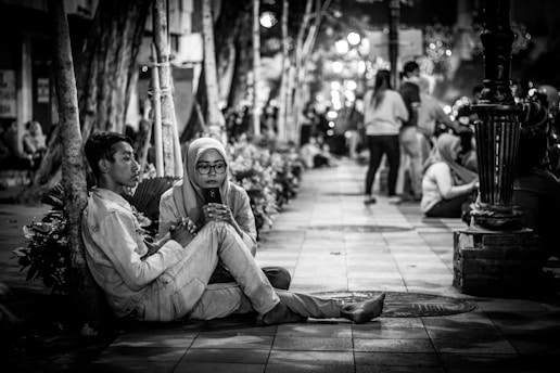 Two people sitting on a tiled pavement, with the woman focused on a mobile phone, possibly engaged in conversation or browsing, while the man sits beside her looking away. The environment is a nighttime urban setting with trees lining the sidewalk and a number of people in the background, some walking and others sitting nearby, giving the scene a relaxed social vibe.