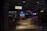 A dimly lit casino with several rows of slot machines, some displaying bright graphics and lights. There are a few people scattered throughout the area, engaging with the machines. The floor is reflective and polished, adding to the ambiance, while the ceiling is dotted with numerous small lights.