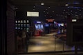 A dimly lit casino with several rows of slot machines, some displaying bright graphics and lights. There are a few people scattered throughout the area, engaging with the machines. The floor is reflective and polished, adding to the ambiance, while the ceiling is dotted with numerous small lights.