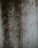 A textured surface displaying dark, uneven patches and speckles, resembling mold or dirt accumulation on a lighter concrete wall.