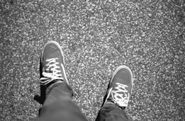 A pair of sneakers with white laces are visible from an overhead view, standing on a textured, gravel-like surface. The sneakers appear worn and are paired with dark jeans.
