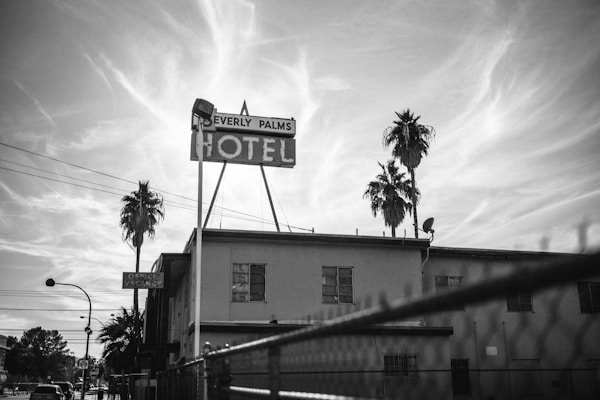 A black-and-white image of a mid-20th-century style motel with a large sign that reads 'Beverly Palms Motel'. Two tall palm trees are silhouetted against a cloudy sky, and a typical suburban street is visible in the background. There are some visible power lines and an old-style satellite dish on the roof.