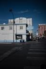 A mid-century style building with a light blue facade features a vintage sign reading 'HOTEL'. It is situated on a city street corner, surrounded by taller modern buildings. A zebra crossing is visible in the foreground, and the lighting suggests it's either early morning or late afternoon.