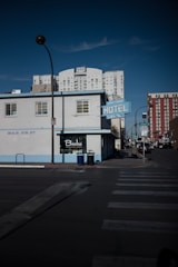 A mid-century style building with a light blue facade features a vintage sign reading 'HOTEL'. It is situated on a city street corner, surrounded by taller modern buildings. A zebra crossing is visible in the foreground, and the lighting suggests it's either early morning or late afternoon.