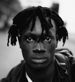 A young person with braided hair looks directly at the camera with a neutral expression. The image is in black and white, creating a strong contrast between the subject's features and the blurred background. The individual wears a casual jacket and carries a backpack over one shoulder, suggesting a street or urban environment.