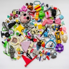 A collection of various colorful plastic toys and objects scattered on a white background. It includes toy cars, dice, hair accessories, miniature figurines, a stuffed animal head, a pink bow with black polka dots, and a photo of a baby in a small frame shaped like an angel. The objects vary in shape, size, and color, creating a chaotic yet playful arrangement.