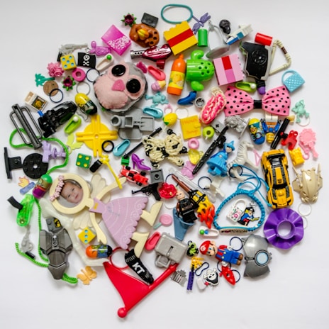 A collection of various colorful plastic toys and objects scattered on a white background. It includes toy cars, dice, hair accessories, miniature figurines, a stuffed animal head, a pink bow with black polka dots, and a photo of a baby in a small frame shaped like an angel. The objects vary in shape, size, and color, creating a chaotic yet playful arrangement.