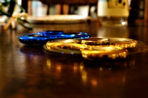 Two fidget spinners, one blue and one gold, resting on a reflective wooden surface. The background is blurred, adding a shallow depth of field effect.