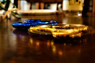 Two fidget spinners, one blue and one gold, resting on a reflective wooden surface. The background is blurred, adding a shallow depth of field effect.