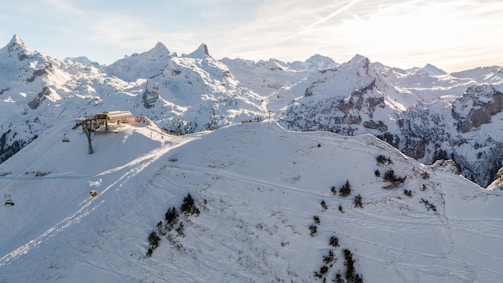 A snowy mountainous landscape with a ski lift station situated on a ridge. The mountains are covered with snow and some pine trees can be seen on the slopes. The sky is mostly clear with a few contrails and the sun is either rising or setting behind the mountains, casting a gentle light over the scene.