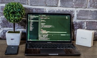 A laptop displaying code is placed on a wooden table, accompanied by a potted plant and a box-like object with inspirational text against a brick wall backdrop. A smartphone is positioned to the left of the laptop.
