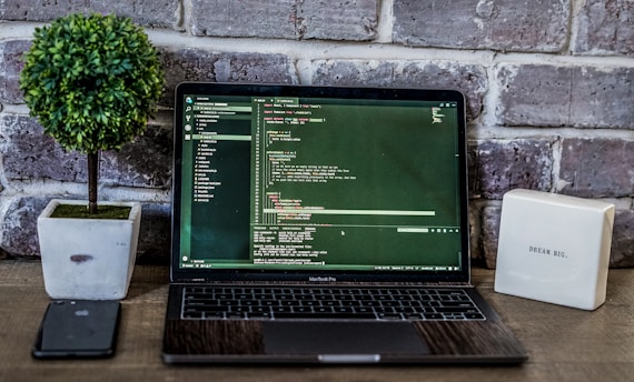 A laptop displaying code is placed on a wooden table, accompanied by a potted plant and a box-like object with inspirational text against a brick wall backdrop. A smartphone is positioned to the left of the laptop.