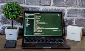 A laptop displaying code is placed on a wooden table, accompanied by a potted plant and a box-like object with inspirational text against a brick wall backdrop. A smartphone is positioned to the left of the laptop.