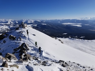 A snow-covered mountain ridge with a clear blue sky overhead. Three hikers equipped with backpacks and trekking poles are ascending the ridge. The vast expanse of mountains and valleys extends into the distance, creating a dramatic landscape.