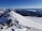 A snow-covered mountain ridge with a clear blue sky overhead. Three hikers equipped with backpacks and trekking poles are ascending the ridge. The vast expanse of mountains and valleys extends into the distance, creating a dramatic landscape.