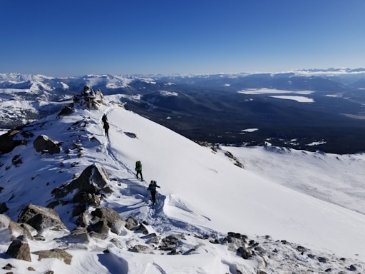 A snow-covered mountain ridge with a clear blue sky overhead. Three hikers equipped with backpacks and trekking poles are ascending the ridge. The vast expanse of mountains and valleys extends into the distance, creating a dramatic landscape.