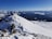 A snow-covered mountain ridge with a clear blue sky overhead. Three hikers equipped with backpacks and trekking poles are ascending the ridge. The vast expanse of mountains and valleys extends into the distance, creating a dramatic landscape.