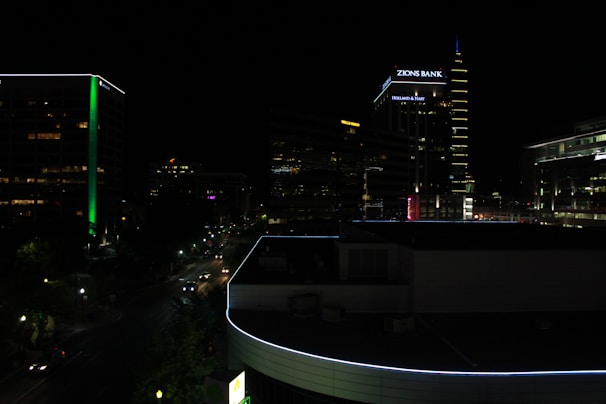 A nighttime cityscape featuring tall buildings with illuminated signs and neon lights. The skyline includes a prominent building displaying 'Zions Bank' and another with a noticeable green vertical stripe. Streets are lit by streetlights and car headlights, with some traffic visible.