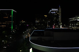 A nighttime cityscape featuring tall buildings with illuminated signs and neon lights. The skyline includes a prominent building displaying 'Zions Bank' and another with a noticeable green vertical stripe. Streets are lit by streetlights and car headlights, with some traffic visible.