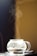 A transparent glass teapot is sitting on a surface with steam gently rising from the spout, indicating it contains hot liquid. The background is softly blurred, with shades of dark blue and warm yellow creating a contrasting ambiance.