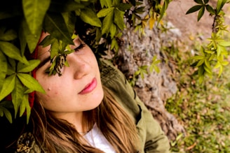 A person is relaxing under a canopy of lush green leaves. The individual appears to be reclining against a tree or rock surface, with their eyes gently closed and face partially obscured by the foliage. The leaves cast dappled shadows, creating a serene and peaceful atmosphere.