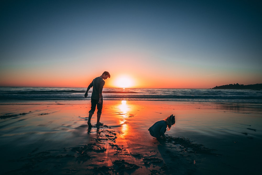 man standing on beach beside girl playing sand during sunset