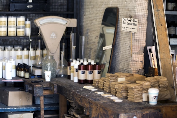 A rustic shop interior with a wooden counter displaying stacks of brown paper-wrapped items, likely soap. Various bottles and jars with labels are arranged on shelves. An antique scale and a mirror are visible in the background. A handwritten sign indicates pricing for soap.