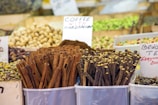 Various spices and nuts are displayed in clear containers at a market stall. Prominent cinnamon sticks and bundles of other spices are accompanied by handwritten signs. The background shows an assortment of other dried goods and nuts.