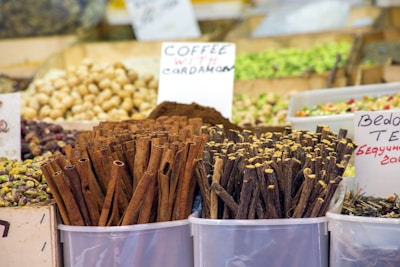 Various spices and nuts are displayed in clear containers at a market stall. Prominent cinnamon sticks and bundles of other spices are accompanied by handwritten signs. The background shows an assortment of other dried goods and nuts.