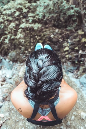 A person with a neatly braided hairstyle is sitting outdoors. The focus is on the back of the head, and the person is wearing a tank top with visible straps. The background shows a blurred natural setting with foliage.