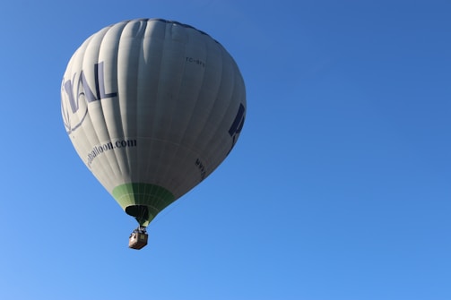 A hot air balloon with a large, patterned envelope floats high in a clear blue sky. The balloon's basket is visible, and text is printed on the side of the envelope.
