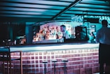 A dimly lit bar with a modern design features a bartender standing behind the counter. The shelves are stocked with various liquor bottles, and a row of empty stools lines the foreground. Glassware is hanging above the bar, and the atmosphere is enhanced by cool blue lighting.