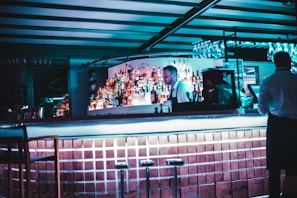 A dimly lit bar with a modern design features a bartender standing behind the counter. The shelves are stocked with various liquor bottles, and a row of empty stools lines the foreground. Glassware is hanging above the bar, and the atmosphere is enhanced by cool blue lighting.