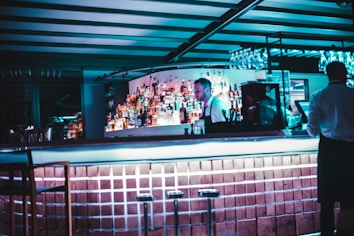 A dimly lit bar with a modern design features a bartender standing behind the counter. The shelves are stocked with various liquor bottles, and a row of empty stools lines the foreground. Glassware is hanging above the bar, and the atmosphere is enhanced by cool blue lighting.