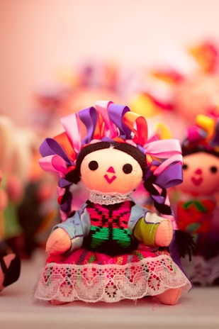 A vibrant, handcrafted doll adorned with colorful ribbons in its braided hair. It wears a traditional outfit featuring intricate embroidery and lace details. The background shows more dolls with similarly colorful attire, softly blurred to emphasize the foreground doll.