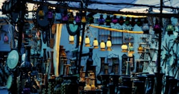 A marketplace stall displaying an array of decorative lanterns, mirrors, and vases. The scene is crowded with various ornamented items, with hanging colored lanterns illuminating the area in warm and cool tones. There are intricate designs and patterns evident in the items, creating an eclectic and vibrant atmosphere.