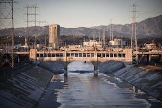 A concrete-lined river channel runs through an urban area flanked by industrial structures and power lines. A bridge with arches spans across the channel, while a tall building towers in the background against a mountain range.