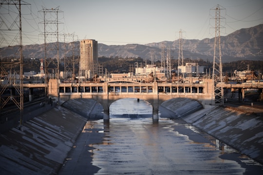 A concrete-lined river channel runs through an urban area flanked by industrial structures and power lines. A bridge with arches spans across the channel, while a tall building towers in the background against a mountain range.