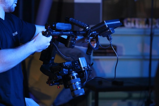 A person is holding a professional video camera rig with various mounted components, including a lens and monitor. The setup is complex and equipped for advanced video production. The background is blurred, suggesting a focus on the equipment.
