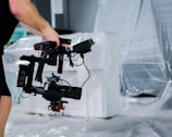 A person holds a professional camera mounted on a stabilizing rig, standing in a space with plastic sheeting covering furniture in the background.