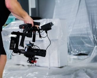 A person holds a professional camera mounted on a stabilizing rig, standing in a space with plastic sheeting covering furniture in the background.